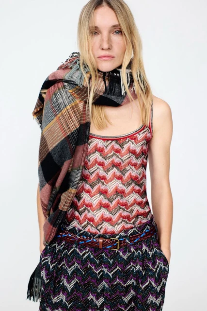 A fashionable woman is wearing a patterned top and pants with a plaid scarf. She has blonde hair and a neutral facial expression.