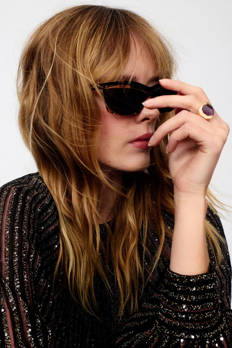 Woman wearing sunglasses and a sparkly top poses against a white backdrop in a medium shot focusing on her face and upper body.