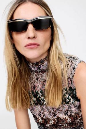 Model wearing dark tortoiseshell sunglasses and a patterned lace top against a white background