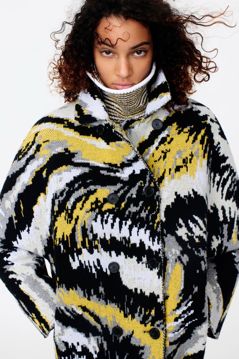 Young adult woman poses in an abstract-patterned, double-breasted coat with a high collar, indoors with a neutral background.