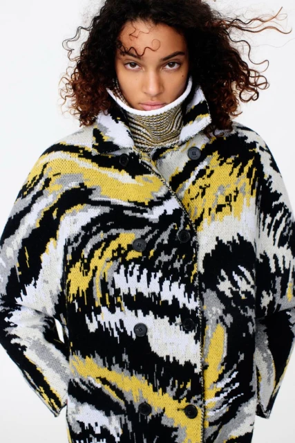 Young adult woman poses in an abstract-patterned, double-breasted coat with a high collar, indoors with a neutral background.