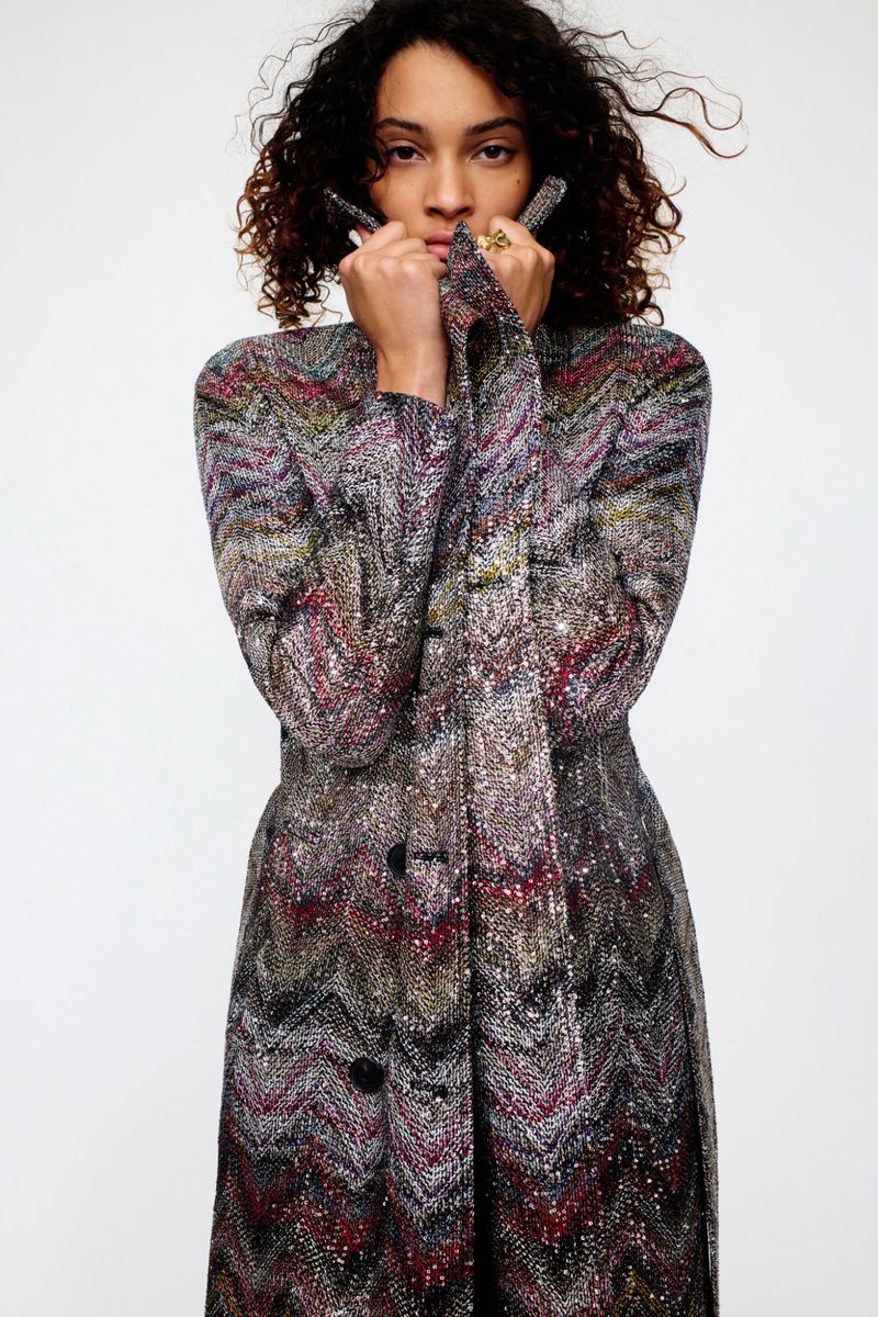 A stylish woman with curly hair wearing a long, patterned coat poses in a studio with a white background, fashion photography.