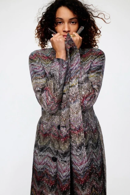 A stylish woman with curly hair wearing a long, patterned coat poses in a studio with a white background, fashion photography.