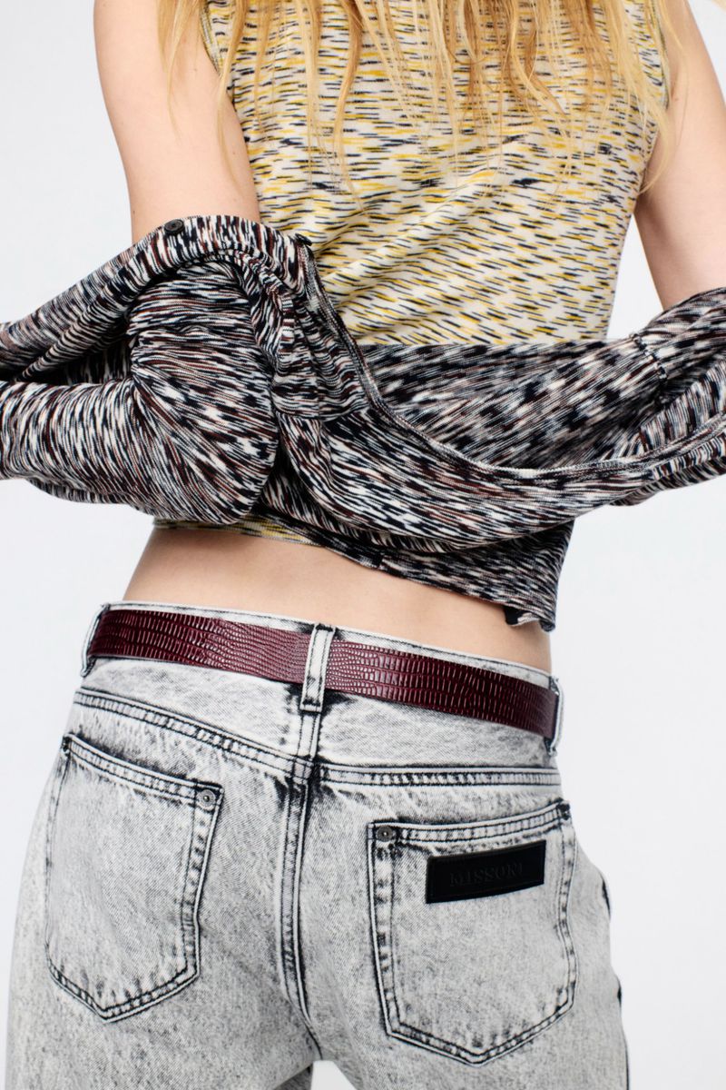 Woman wearing a yellow and grey patterned top, grey jeans, and a maroon belt is seen from the back in a studio setting.