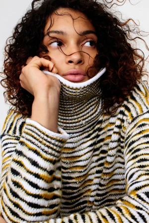 Model with curly dark hair wearing a striped turtleneck sweater against a white background