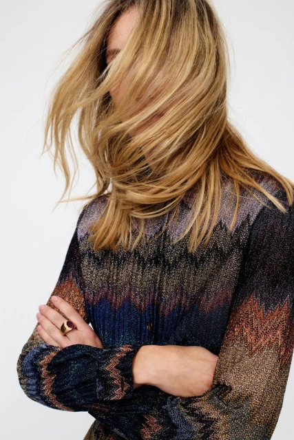 Fashion shot of a woman with blonde hair covering her face, wearing a chevron-patterned knit top and a gold ring, against a white backdrop.