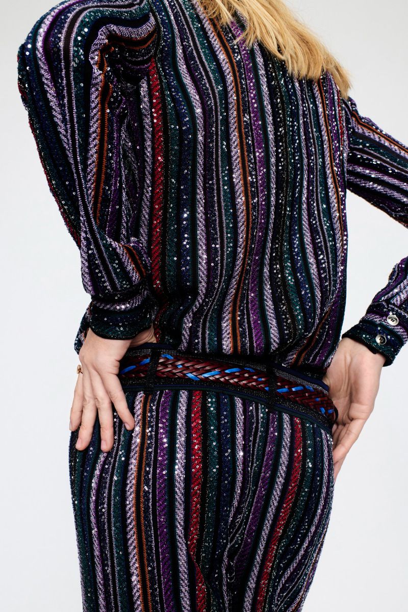 Back view of an adult female wearing a multicolored striped sequin outfit against a white background in a fashion studio setting.