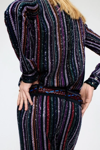 Back view of an adult female wearing a multicolored striped sequin outfit against a white background in a fashion studio setting.