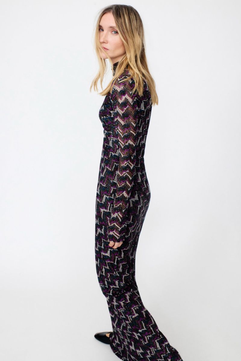 Blonde woman in a black patterned dress posing in a studio. The dress has long sleeves and a high neck. The background is white.