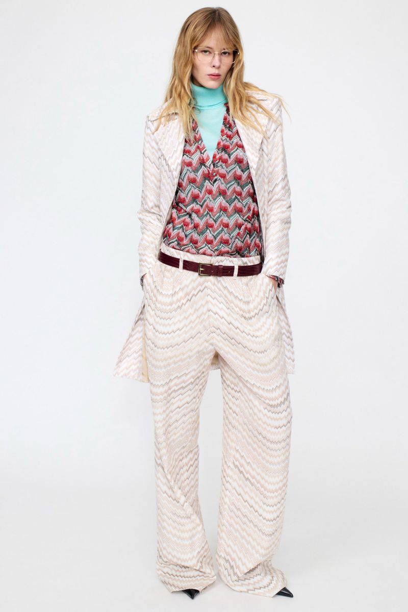 Model standing against a white backdrop wearing a patterned cream coat, matching trousers, and a blue turtleneck.