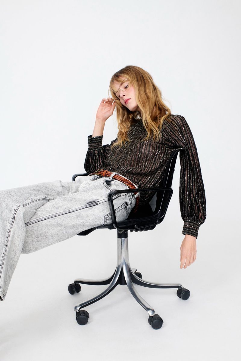 Adult female model in long sleeve, black and gold top and grey denim sitting on a black office chair against a white backdrop.