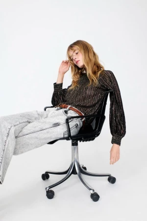 Adult female model in long sleeve, black and gold top and grey denim sitting on a black office chair against a white backdrop.