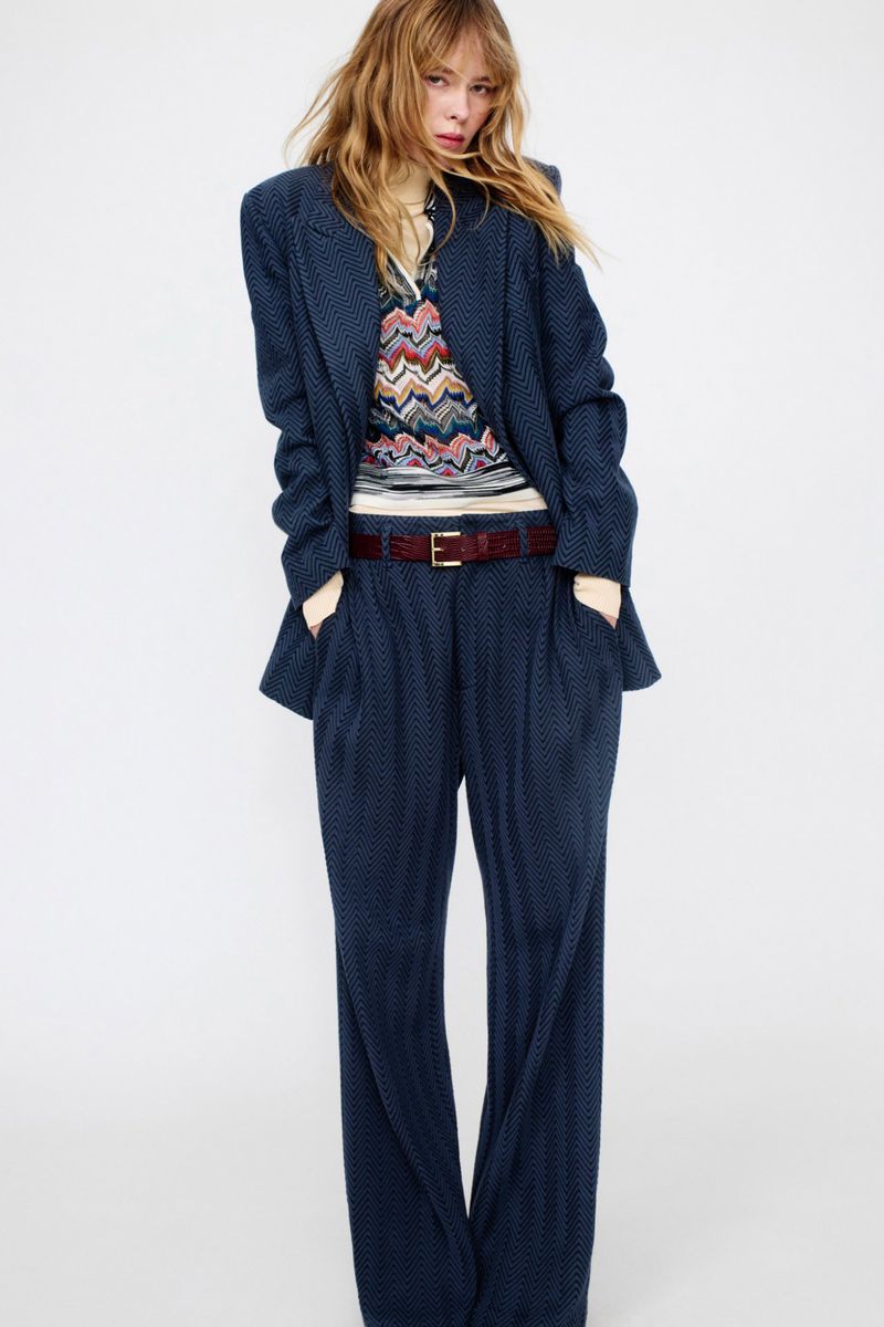 Adult female fashion model poses in a patterned navy blue blazer and trousers with a colorful top against a neutral backdrop indoors.