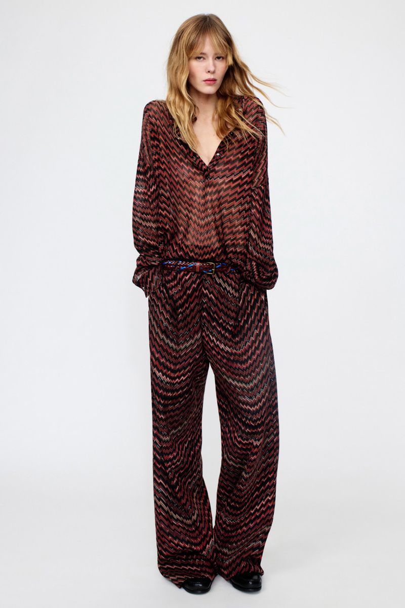 Fashionable woman poses in a long-sleeved blouse and wide-leg trousers with a zigzag pattern against a clean, bright background.