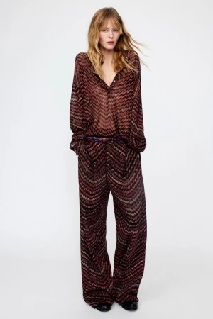 Fashionable woman poses in a long-sleeved blouse and wide-leg trousers with a zigzag pattern against a clean, bright background.