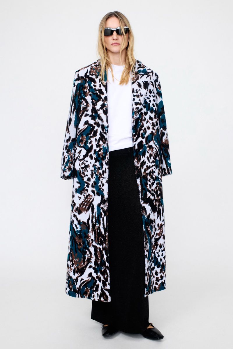 A woman with long hair stands indoors wearing sunglasses, a long animal-print coat, a white shirt, and a black skirt against a white background.