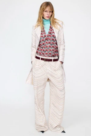 Model standing against a white backdrop wearing a patterned cream coat, matching trousers, and a blue turtleneck.