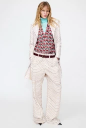 Model standing against a white backdrop wearing a patterned cream coat, matching trousers, and a blue turtleneck.