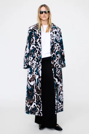 Model wearing patterned coat and black trousers standing against a white seamless background