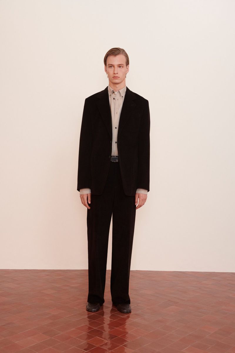 Blond male model in a black suit stands against a light wall on a red tile floor in a medium studio shot promoting fashion.