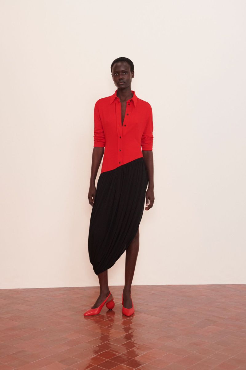 A dark-skinned woman in a red shirt and black skirt stands in a minimalist setting, modeling fashion with red shoes. The shot is full length.