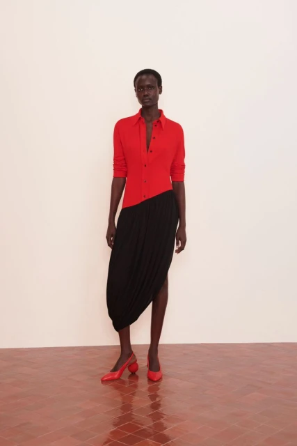 A dark-skinned woman in a red shirt and black skirt stands in a minimalist setting, modeling fashion with red shoes. The shot is full length.