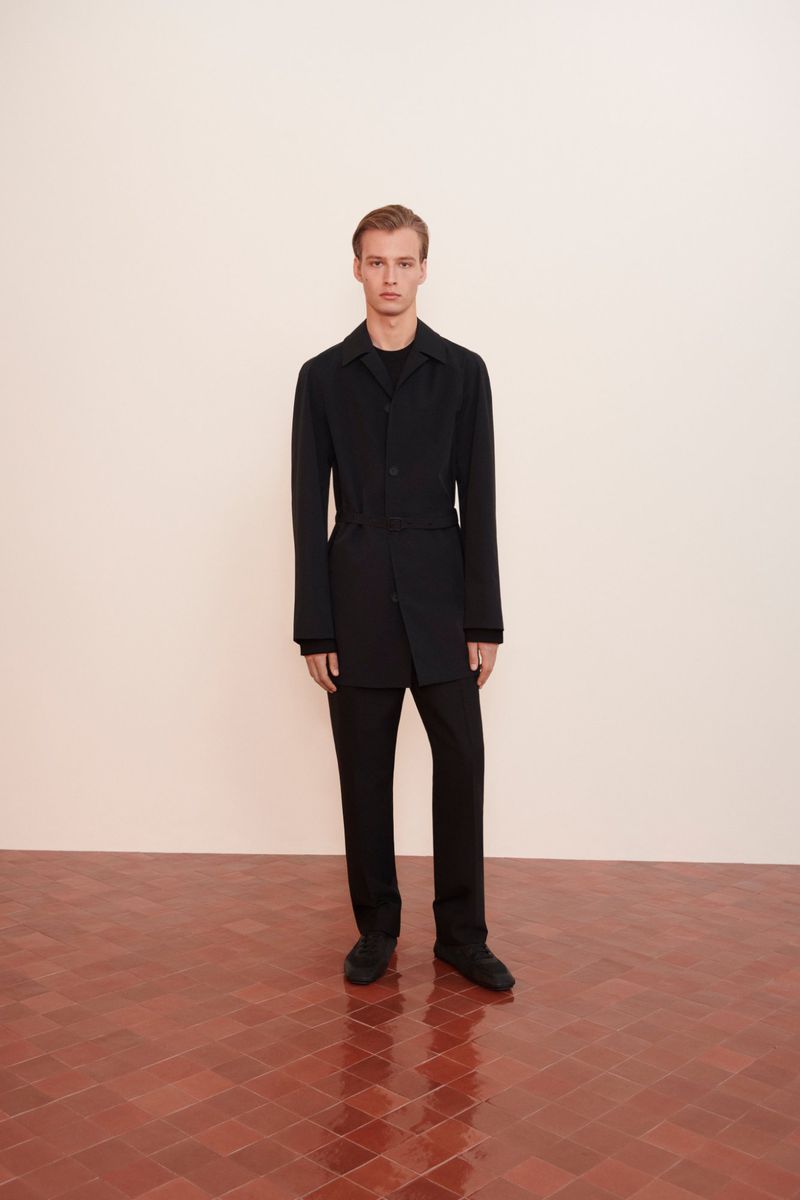 A blond male model stands in an interior wearing a black jacket and trousers, posing against a pale peach wall and a brick-tiled floor.
