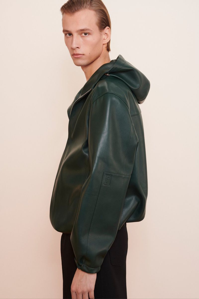 Side-angle shot of a male model in a green leather hooded jacket and black pants, posing against a neutral background in a studio setting.