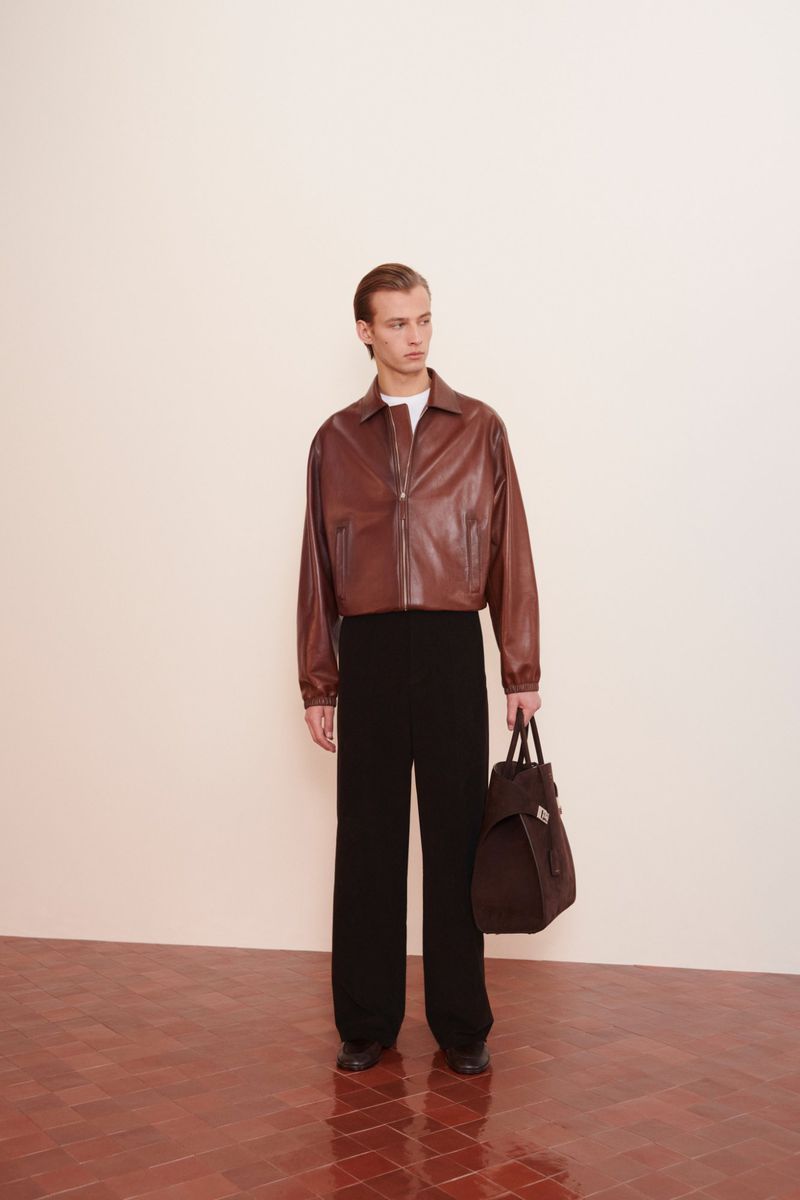 Male model in leather jacket, trousers, and bag in studio. Neutral colors and clean lines. Fashion editorial, modern menswear style.