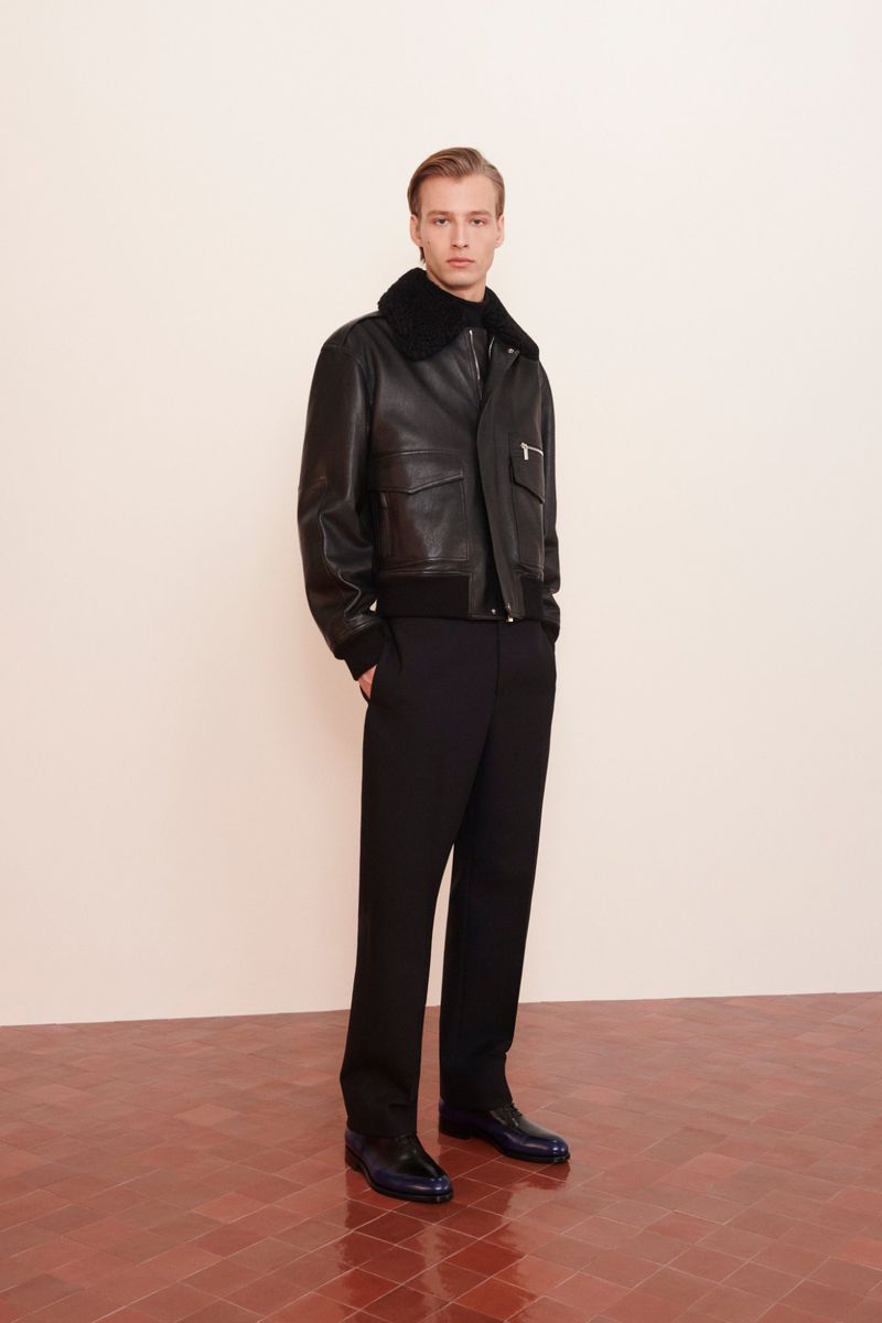 Man in black leather bomber jacket with fur collar, black shirt, and dark pants poses in a room with a red and white tiled floor.