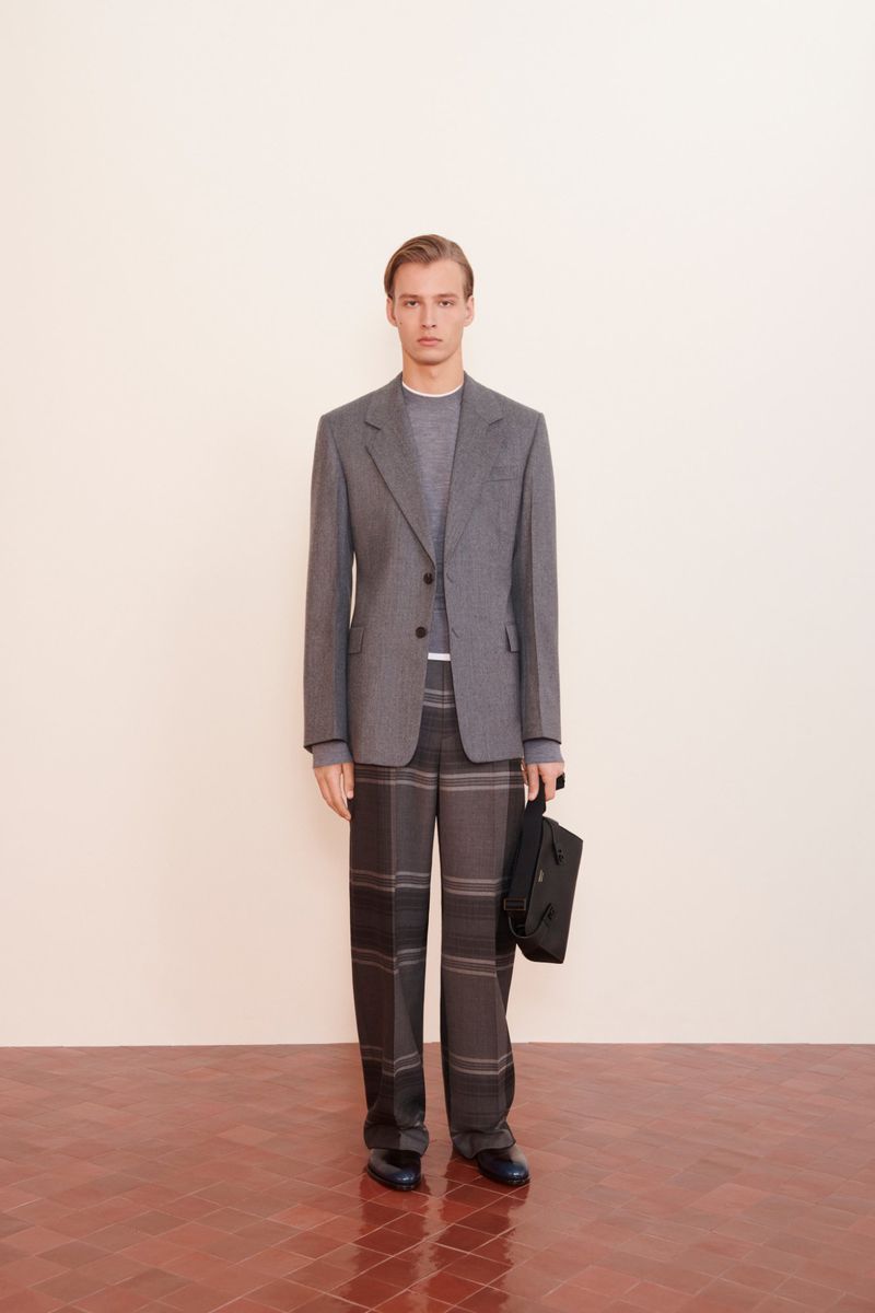A fair-skinned adult male model poses in a grey blazer and plaid trousers, indoors, holding a black rectangular bag, on a brick floor.