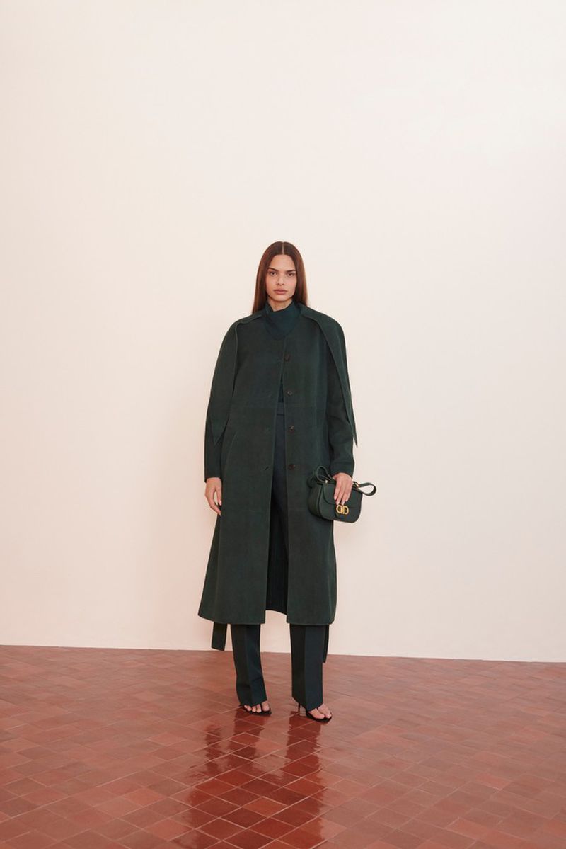 A brunette woman poses in a long dark green coat and matching pants, holding a black purse, against a cream background and terra cotta floor.