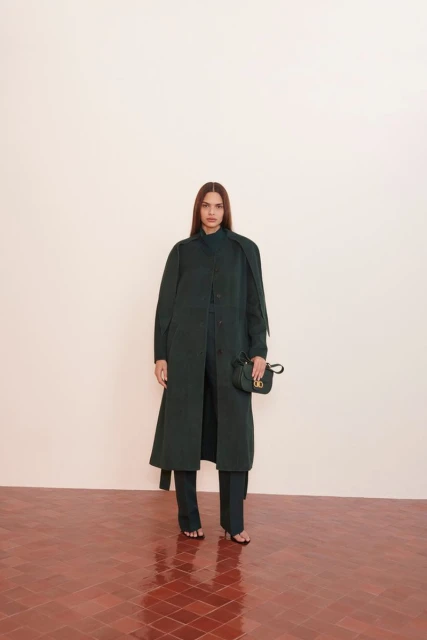 A brunette woman poses in a long dark green coat and matching pants, holding a black purse, against a cream background and terra cotta floor.