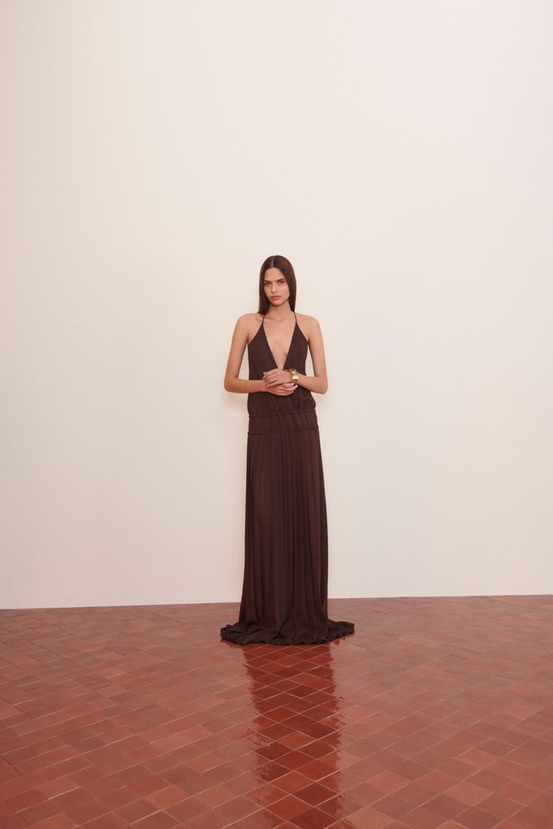 An adult female wearing a dark brown halter dress is standing indoors on a red tile floor, posing against a white wall.