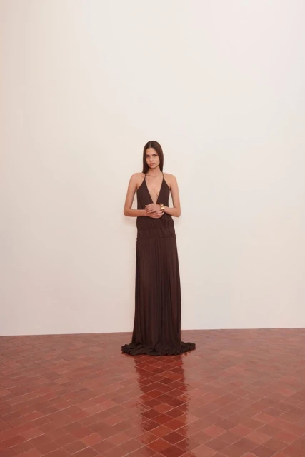 An adult female wearing a dark brown halter dress is standing indoors on a red tile floor, posing against a white wall.