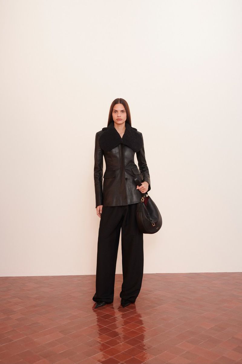 Model wearing a black leather jacket with a large collar and wide black trousers, carrying a black shoulder bag.