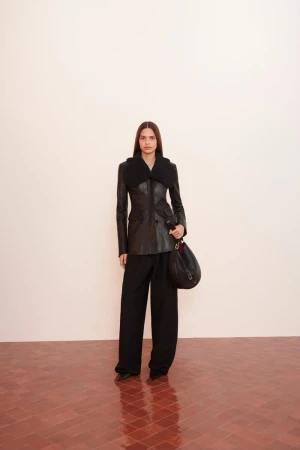 Model wearing a black leather jacket with a large collar and wide black trousers, carrying a black shoulder bag.
