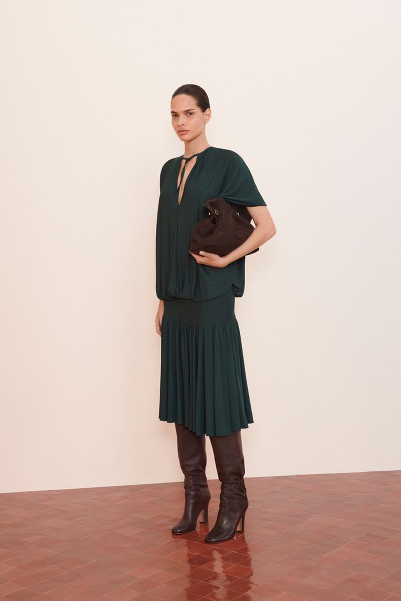 A woman in a dark green dress and boots holds a handbag against a white wall and red herringbone floor, fashion editorial.