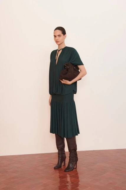 A woman in a dark green dress and boots holds a handbag against a white wall and red herringbone floor, fashion editorial.