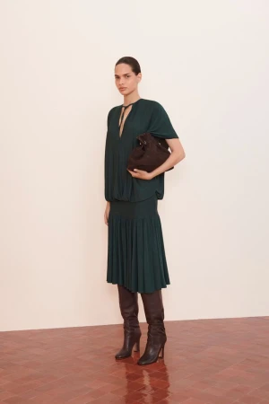 A woman in a dark green dress and boots holds a handbag against a white wall and red herringbone floor, fashion editorial.