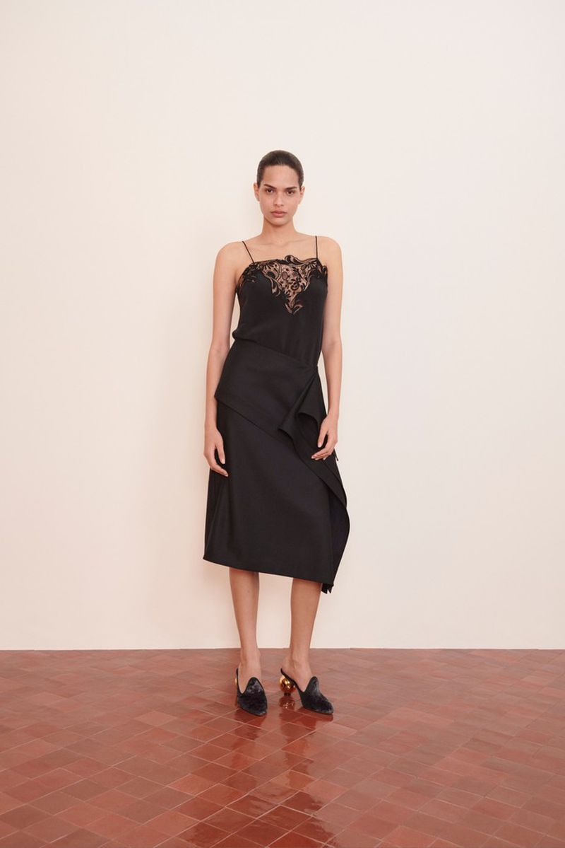 A woman stands in a photo studio modeling a black lace top, black skirt, and black heels against a white wall and terracotta floor.