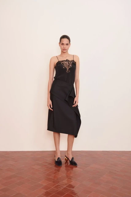 A woman stands in a photo studio modeling a black lace top, black skirt, and black heels against a white wall and terracotta floor.