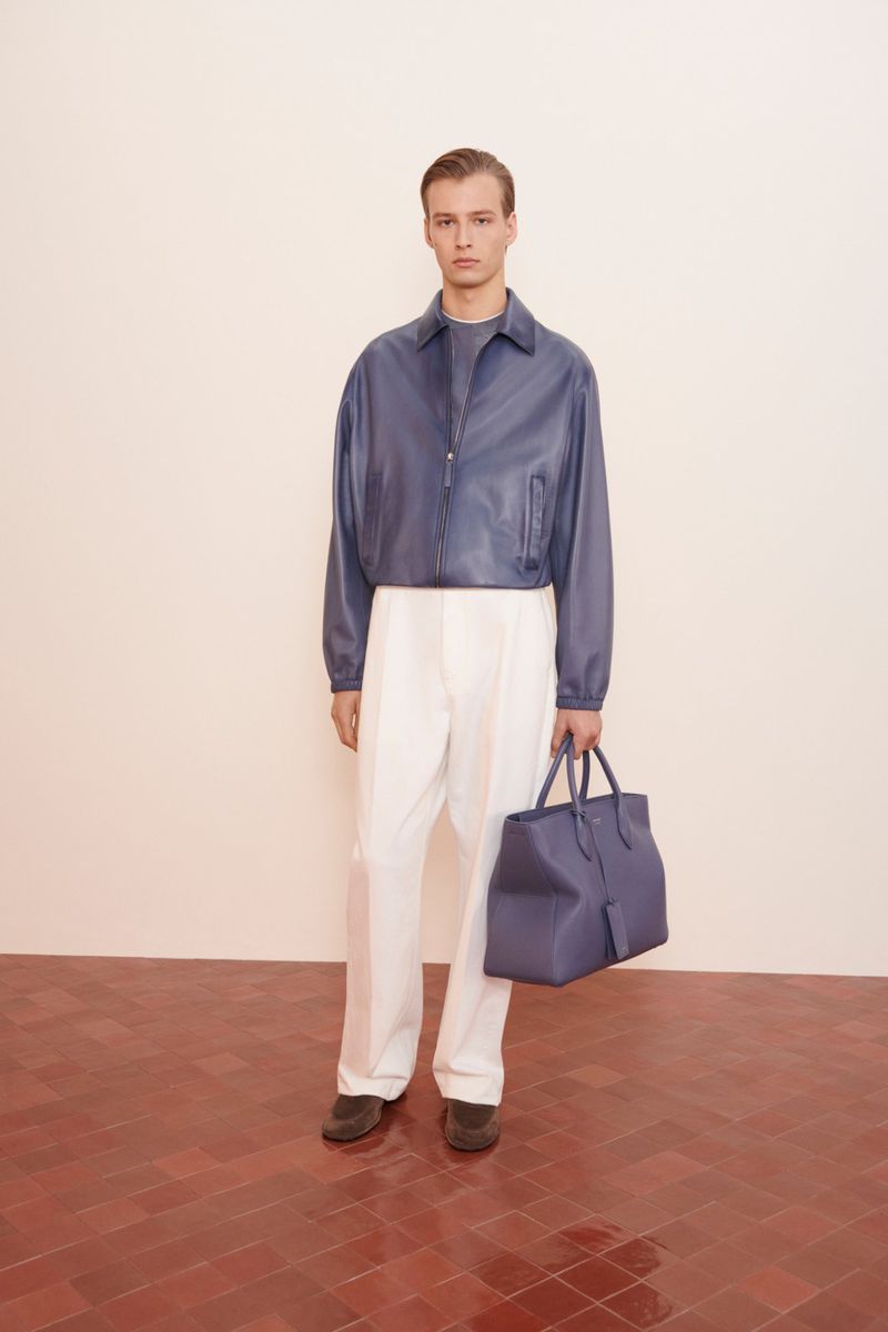 A male fashion model poses in a photo studio wearing a blue leather jacket and white pants, holding a blue leather tote bag.
