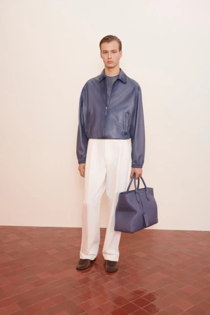 A male fashion model poses in a photo studio wearing a blue leather jacket and white pants, holding a blue leather tote bag.