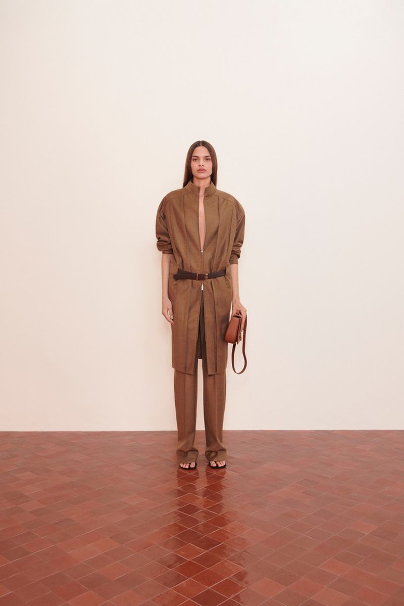 Fashionable woman in brown pantsuit with belt and purse, posing indoors with a neutral background and terracotta tile floor.