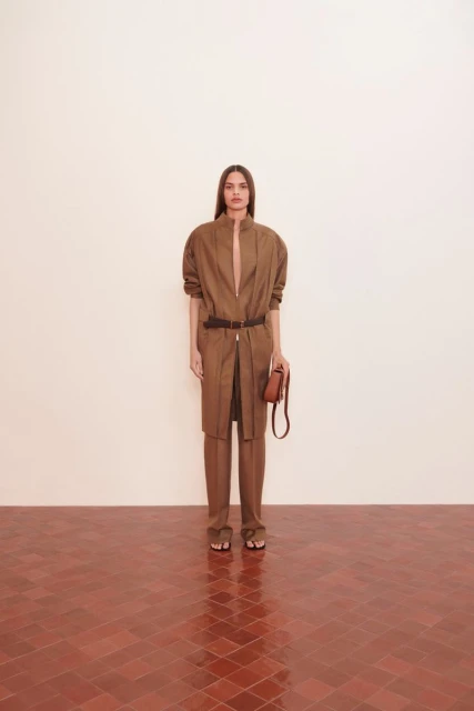 Fashionable woman in brown pantsuit with belt and purse, posing indoors with a neutral background and terracotta tile floor.