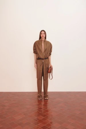 Fashionable woman in brown pantsuit with belt and purse, posing indoors with a neutral background and terracotta tile floor.