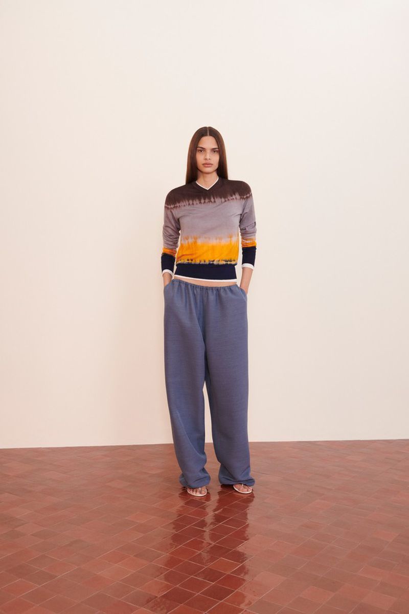 A young woman in a tie-dye top and blue sweatpants stands in front of a neutral wall on a red-tiled floor, full studio shot.