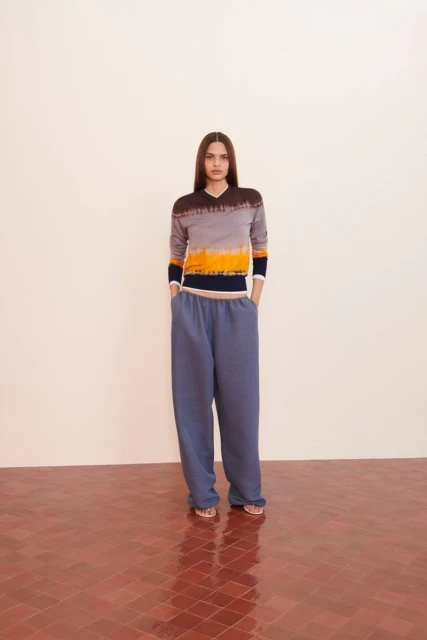 A young woman in a tie-dye top and blue sweatpants stands in front of a neutral wall on a red-tiled floor, full studio shot.