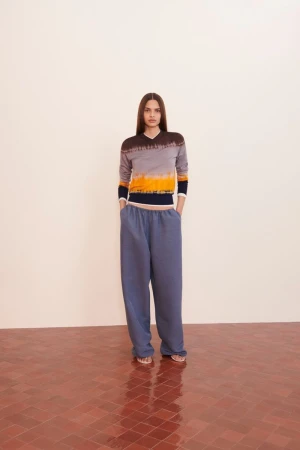 A young woman in a tie-dye top and blue sweatpants stands in front of a neutral wall on a red-tiled floor, full studio shot.